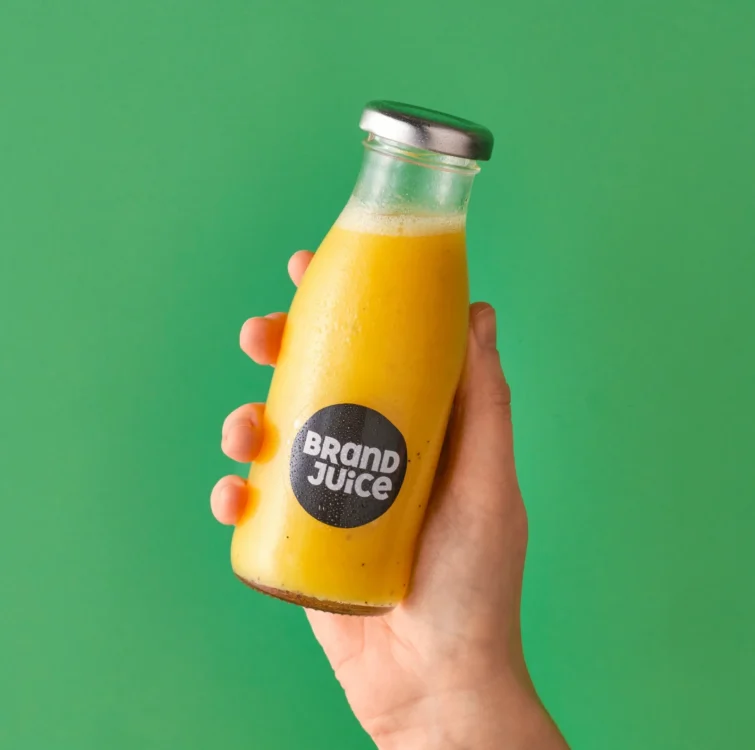 A hand holding a glass bottle of yellow coloured juice on a green background