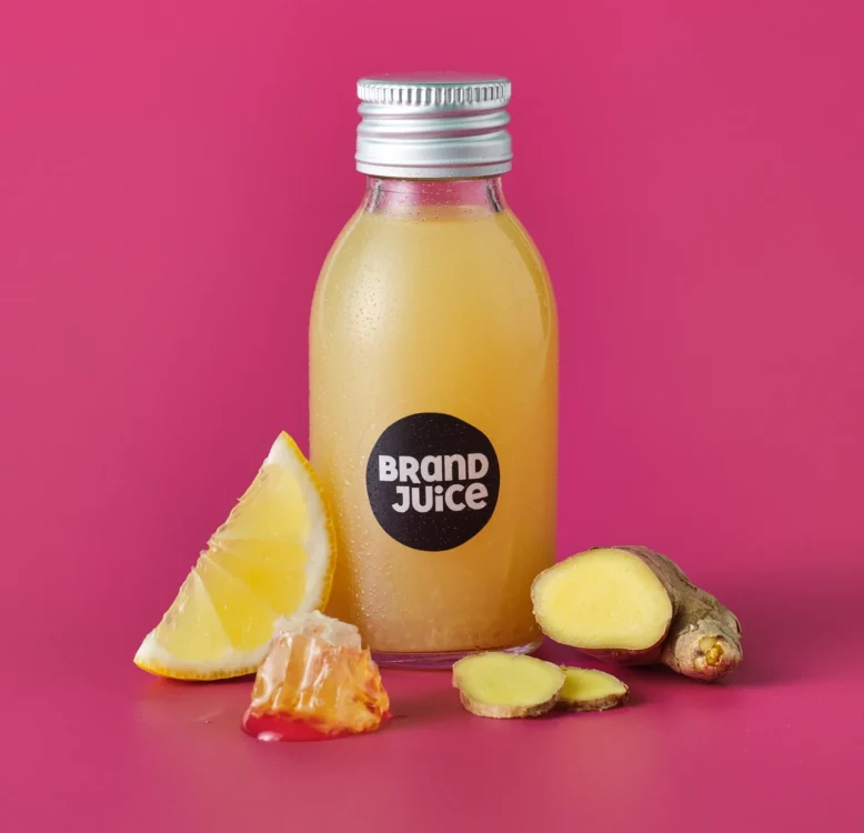 A small bottle of branded juice shot with ginger, orange and honey ingredients next to it