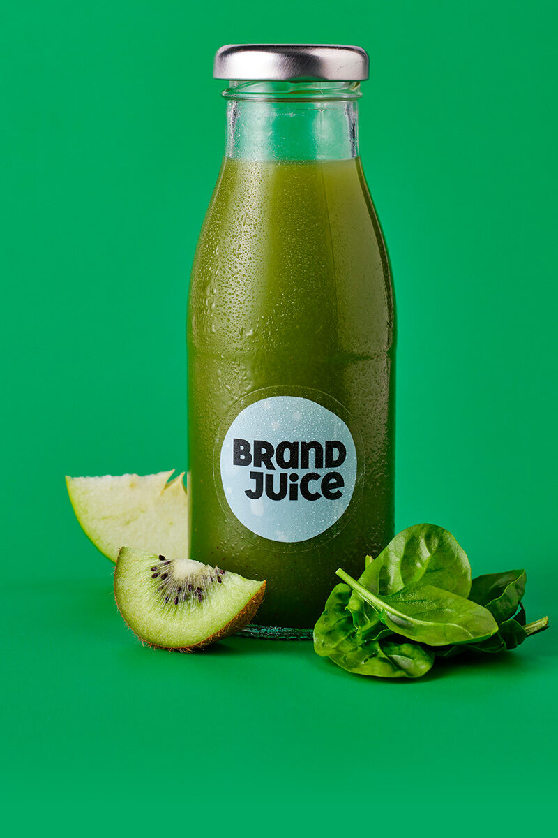 A bottle of green juice with a clear branded label. The ingredients of the juice are next to the bottle