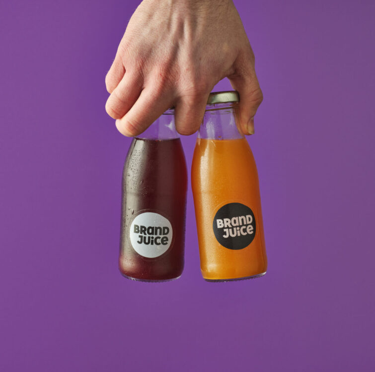 A hand holding two bottles of juice with custom branded labels on a purple background